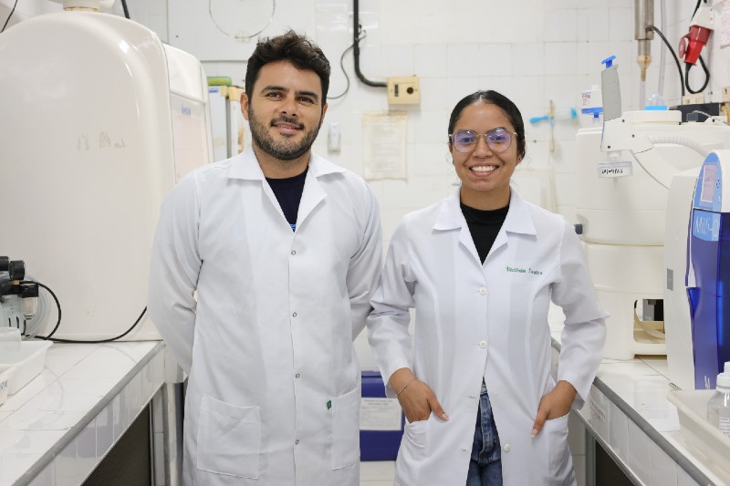 Duas pessoas usando jalecos brancos posam em um laboratório, ao lado de equipamentos científicos e bancadas de trabalho