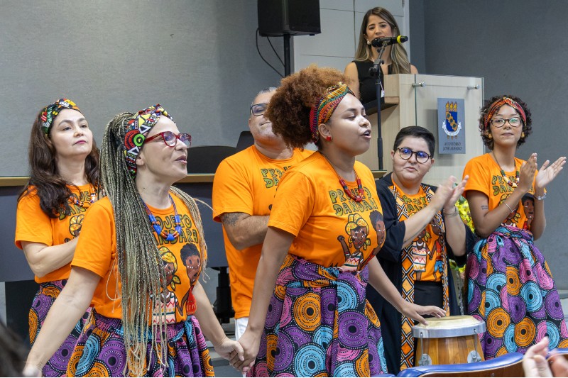 Integrantes do coletivo Dandara cantam e dançam dentro do auditório da UFC, mulheres vestem camiseta laranja e saias coloridas
