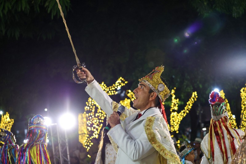 Homem caracterizado como rei segura uma espada e um microfone durante uma apresenta&ccedil;&atilde;o noturna, cercado por dan&ccedil;arinos fantasiados e ilumina&ccedil;&atilde;o decorativa