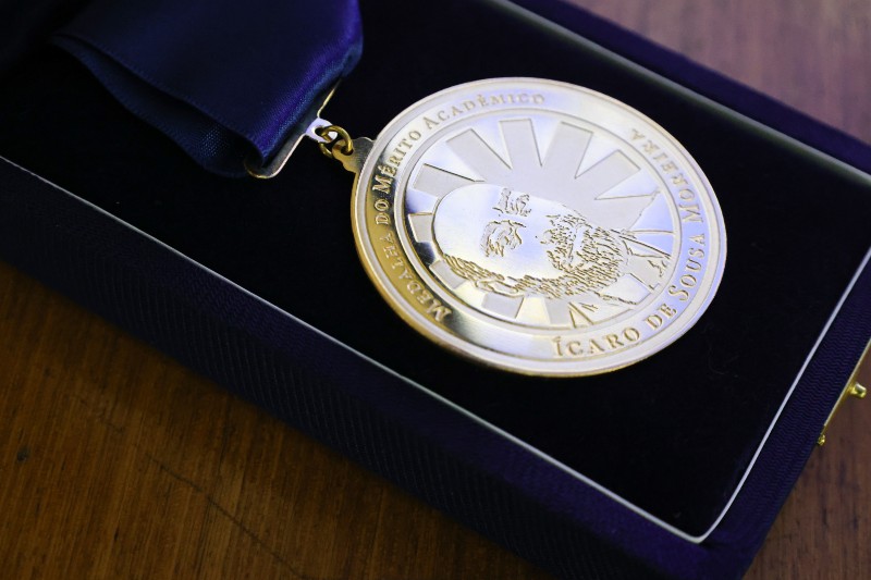 Medalha acadêmica dourada, com imagem em relevo de uma figura humana e inscrições, acomodada em estojo azul-escuro sobre uma superfície de madeira