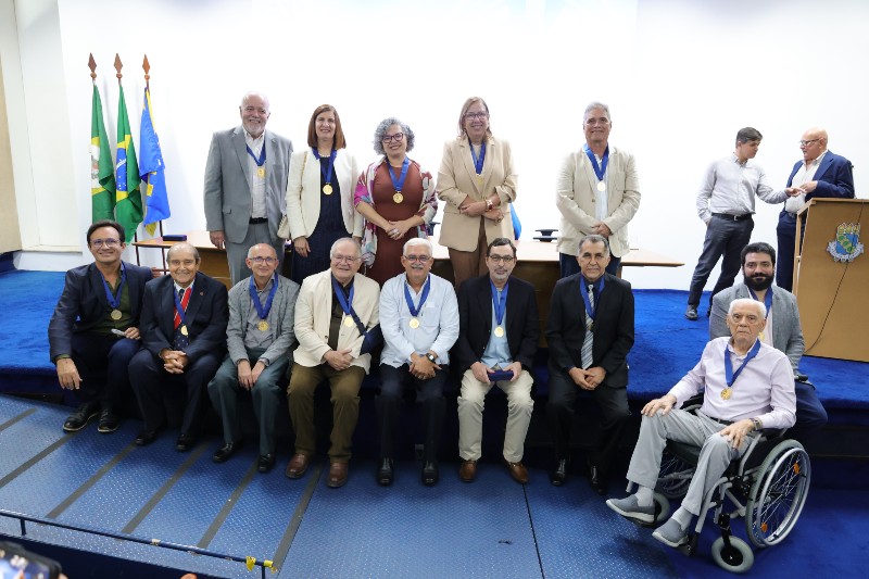 Foto de grupo com 14 pessoas usando medalhas, reunidas em um audit&oacute;rio diante das bandeiras do Brasil e do Cear&aacute;. Ao fundo, h&aacute; uma mesa e um p&uacute;lpito com o bras&atilde;o da UFC