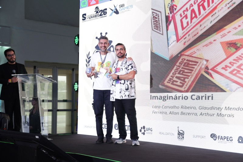 Dois homens recebendo pr&ecirc;mio em evento de games, posando para foto em cima do palco
