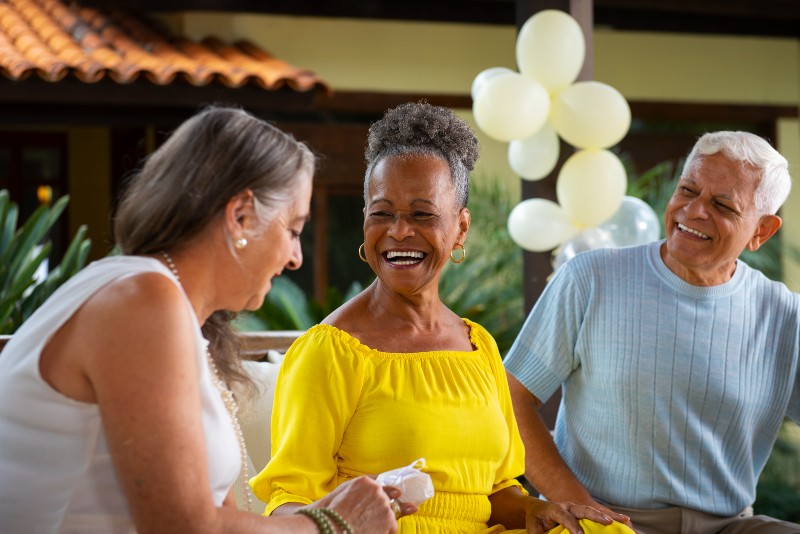 Tr&ecirc;s pessoas idosas conversam e sorriem durante uma comemora&ccedil;&atilde;o. Ao centro, uma mulher negra veste amarelo e segura um presente. Ao fundo, h&aacute; bal&otilde;es brancos e um ambiente externo decorado.