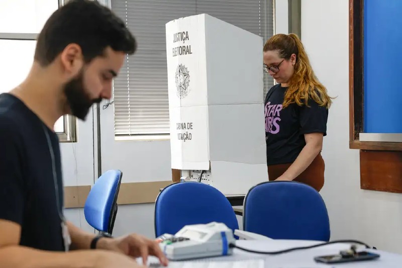 Em primeiro plano, mes&aacute;rio trabalhando e, ao fundo, mulher votando em cabine eleitoral