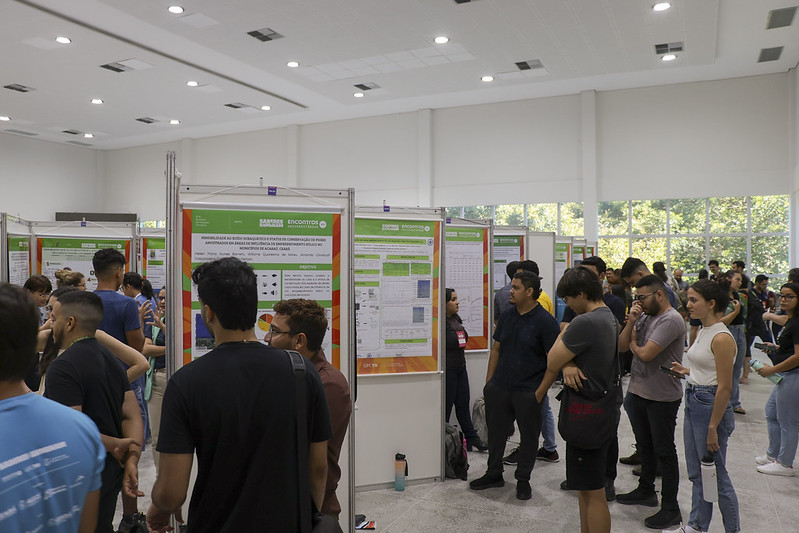 V&aacute;rios alunos em p&eacute; observando p&ocirc;steres cient&iacute;ficos nos Encontros Universit&aacute;rios
