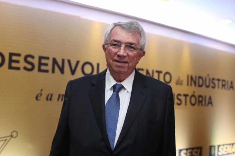 Roberto Mac&ecirc;do aparece em p&eacute;, usando terno preto, camisa branca e gravata azul. Ele sorri discretamente diante de um painel dourado com inscri&ccedil;&otilde;es sobre o desenvolvimento industrial.