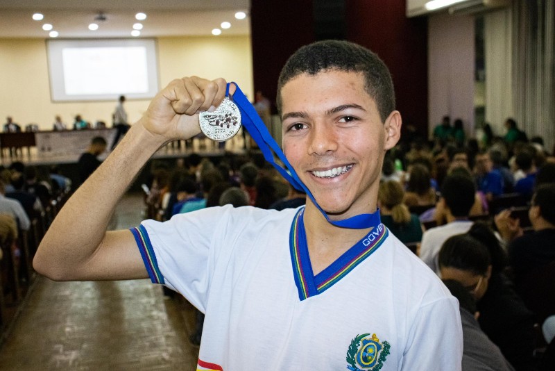 Jovem com farda e uma escola segurando uma medalha conquistada em olimp&iacute;ada do conhecimento
